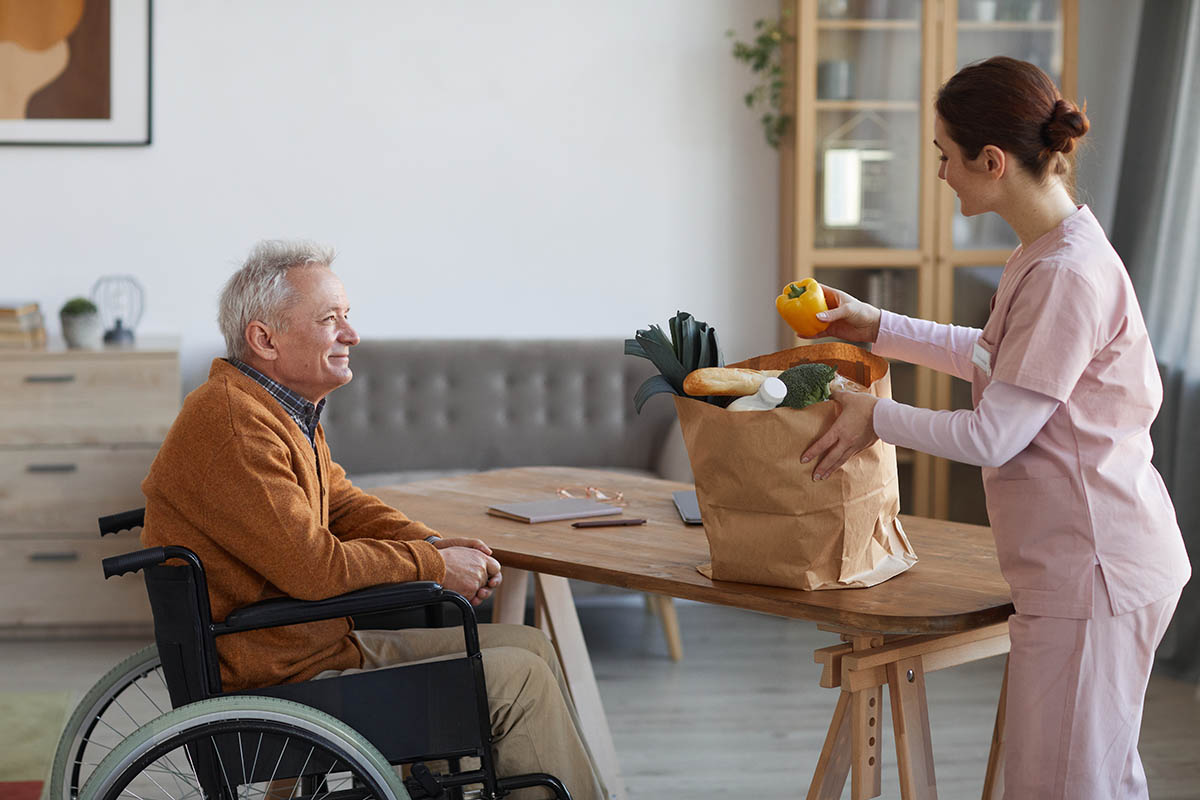 Ein lächelnder Senior im Rollstuhl erhält einen Einkauf, den eine Haushaltshilfe zu ihm nach Hause bringt.